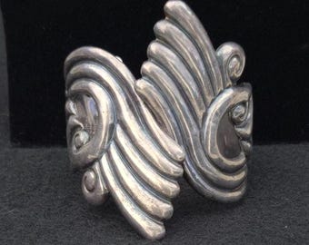 May include: A wide silver cuff bracelet featuring a polished metallic finish with raised, swirling, and fan-like embossed patterns that curve across the surface. The design consists of symmetrical, flowing lines that create a textured appearance.