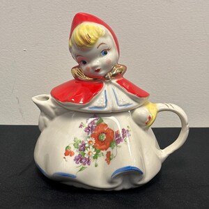 May include: A vintage ceramic teapot shaped like a girl wearing a red hooded dress with floral accents. The teapot has a spout and a handle.