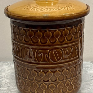 May include: A brown ceramic jar with a lid, featuring a raised design of repeating teardrop shapes. The jar has the word "Rumtopf" written on the lid and the word "Rumtopf" written on the side of the jar.