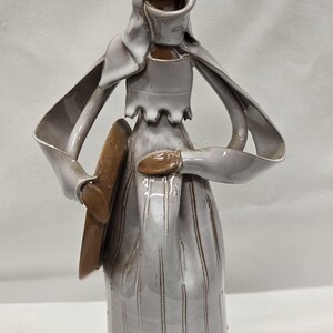 May include: A ceramic sculpture of a figure in a long dress and a head covering. The figure is primarily light gray with brown accents. The sculpture has a unique, abstract design and is likely a decorative art piece.