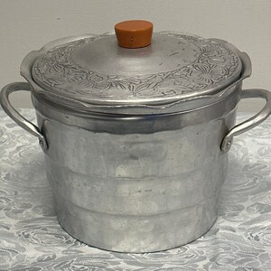 May include: A silver aluminum ice bucket with a floral embossed lid and an orange plastic knob. The bucket has two handles and is sitting on a white and gray patterned surface.