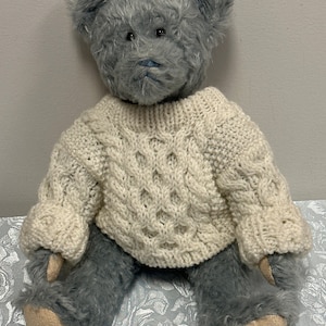 May include: A gray teddy bear wearing a white cable knit sweater. The bear is sitting on a white floral patterned surface.
