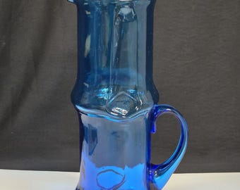 May include: A tall, cobalt blue glass pitcher featuring a cylindrical body with a distinct pinched or indented midsection, a wide pouring spout, and a curved handle attached to the side. The glass has a smooth, translucent finish throughout.