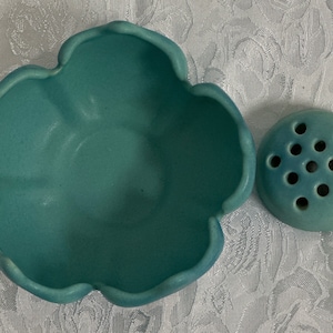 May include: A teal blue ceramic flower frog with a round base and multiple holes for flower stems. The frog is paired with a matching teal blue ceramic bowl with a scalloped edge.