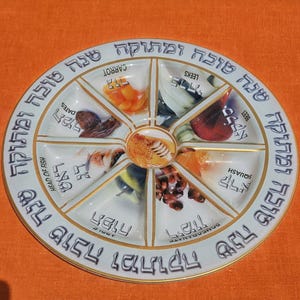 May include: A white ceramic plate with a gold rim, divided into segments, each containing a different food item. The plate has Hebrew text around the edge and labels for each food item. The plate is on an orange surface.