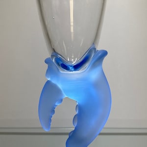 May include: A clear glass goblet with a frosted blue hand-shaped base. The hand is holding the goblet up.