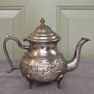May include: A silver teapot with an ornate design and three legs. The teapot has a lid with a small knob on top.