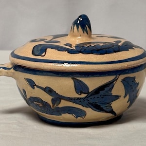 May include: A small, white ceramic bowl with a lid, decorated with blue floral and bird designs. The bowl has two handles on the sides.