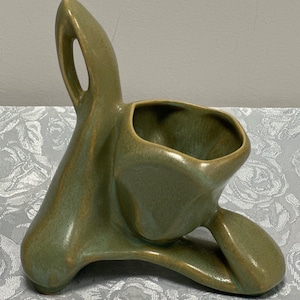 May include: A green ceramic planter with an abstract, organic shape. The planter has a single handle and a wide, open top.