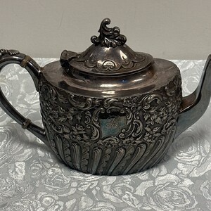 May include: A silver teapot with an ornate design and a lid with a finial. The teapot has a handle and a spout. The teapot is decorated with a floral pattern and a monogram.