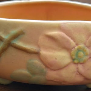 May include: A pale pink ceramic bowl with a raised floral design. The bowl has three feet and features a large pink flower with a yellow center and green leaves.