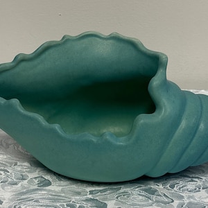 May include: A turquoise ceramic seashell planter with a textured surface. The planter is shaped like a conch shell with a wide opening and a spiral tail.