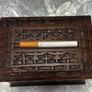 May include: A dark brown wooden box with intricate carvings. The box has a slot for a cigarette, and a cigarette is placed in the slot.