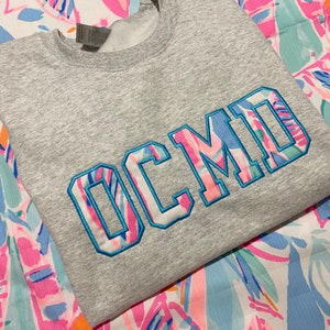 May include: A heather gray sweatshirt with the embroidered letters "OCMD" in a colorful floral pattern. The letters are outlined in blue stitching. The sweatshirt is displayed on a patterned fabric with pink, blue, and white designs.