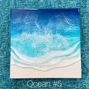 May include: Square artwork depicting an ocean scene. The top portion shows deep blue water transitioning to turquoise with white foamy waves. The bottom features a sandy beach. The text "Ocean #5" is at the bottom.