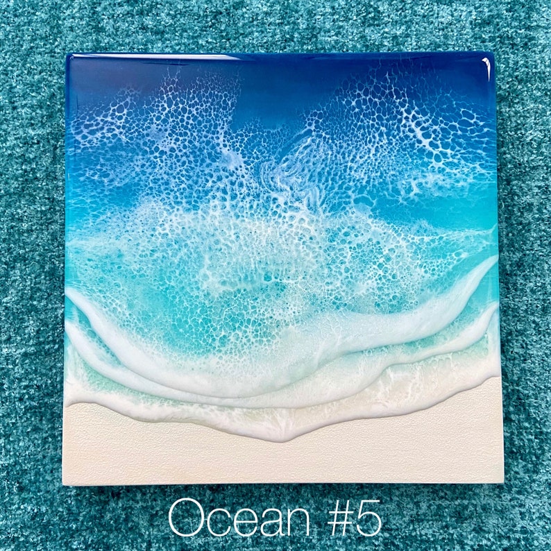 May include: Square artwork depicting an ocean scene. The top portion shows deep blue water transitioning to turquoise with white foamy waves. The bottom features a sandy beach. The text "Ocean #5" is at the bottom.
