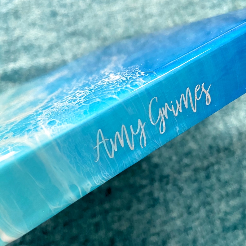 May include: A rectangular art piece with a blue and white ocean wave design. The artwork features the name "Amy Grimes" in a white script font on the side. The piece has a glossy finish and is angled towards the viewer.