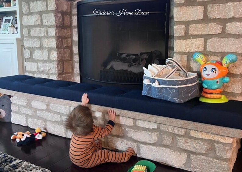 Puede incluir: Un coj&iacute;n azul marino cubre la repisa de una chimenea de piedra. Una cesta estampada en gris y blanco con asas est&aacute; sobre el coj&iacute;n, junto a un juguete interactivo colorido. Un ni&ntilde;o con pijama a rayas naranjas se estira hacia la repisa.