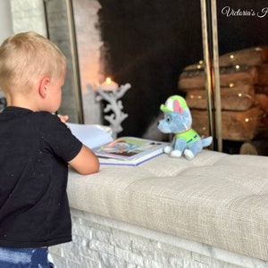 Puede incluir: Un ni&ntilde;o leyendo un libro en un banco tapizado beige. Un peluche azul y verde con forma de cachorro est&aacute; cerca. La escena est&aacute; ambientada frente a una chimenea con velas decorativas y el texto "Victoria's Home".