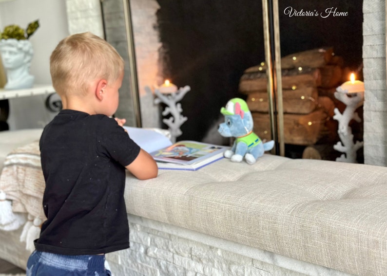 Puede incluir: Un ni&ntilde;o leyendo un libro en un banco tapizado beige. Un peluche azul y verde con forma de cachorro est&aacute; cerca. La escena est&aacute; ambientada frente a una chimenea con velas decorativas y el texto "Victoria's Home".