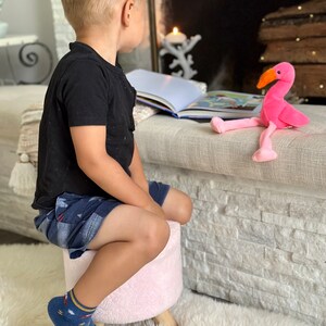 Puede incluir: Un juguete de flamenco de peluche rosa con pico naranja y patas largas est&aacute; cerca de un libro abierto. Un ni&ntilde;o peque&ntilde;o con una camisa negra, pantalones cortos de mezclilla y calcetines azules con un patr&oacute;n est&aacute; sentado en un taburete rosa.