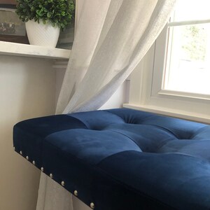 K&ouml;nnte beinhalten: Eine marineblaue Samtbank mit Knopfheftung und silbernen Ziernieten. Die Bank befindet sich in der N&auml;he eines Fensters mit wei&szlig;en, transparenten Vorh&auml;ngen und einer dekorativen Pflanze in einem wei&szlig;en Topf.