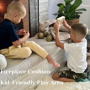 May include: Two young children playing on a large, neutral-toned cushion in front of a fireplace. A decorative white pumpkin and several acorn-shaped objects are visible. The text "Fireplace Cushion Kid-Friendly Play Area" is displayed.