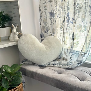 K&ouml;nnte beinhalten: Ein flauschiges, wei&szlig;es herzf&ouml;rmiges Kissen liegt auf einem grauen Fensterbanksitz. Das Fenster wird von floral gemusterten Vorh&auml;ngen eingerahmt. Eine Keramik-Hasenfigur und Topfpflanzen sind ebenfalls zu sehen.