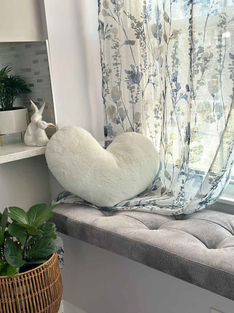 K&ouml;nnte beinhalten: Ein flauschiges, wei&szlig;es herzf&ouml;rmiges Kissen liegt auf einem grauen Fensterbanksitz. Das Fenster wird von floral gemusterten Vorh&auml;ngen eingerahmt. Eine Keramik-Hasenfigur und Topfpflanzen sind ebenfalls zu sehen.