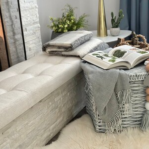 Puede incluir: Una escena interior acogedora con un banco acolchado, una manta a rayas grises y blancas y un libro abierto. Una cesta gris tejida contiene un lanzamiento gris con flecos. Un oso de peluche blanco y cuentas decorativas.