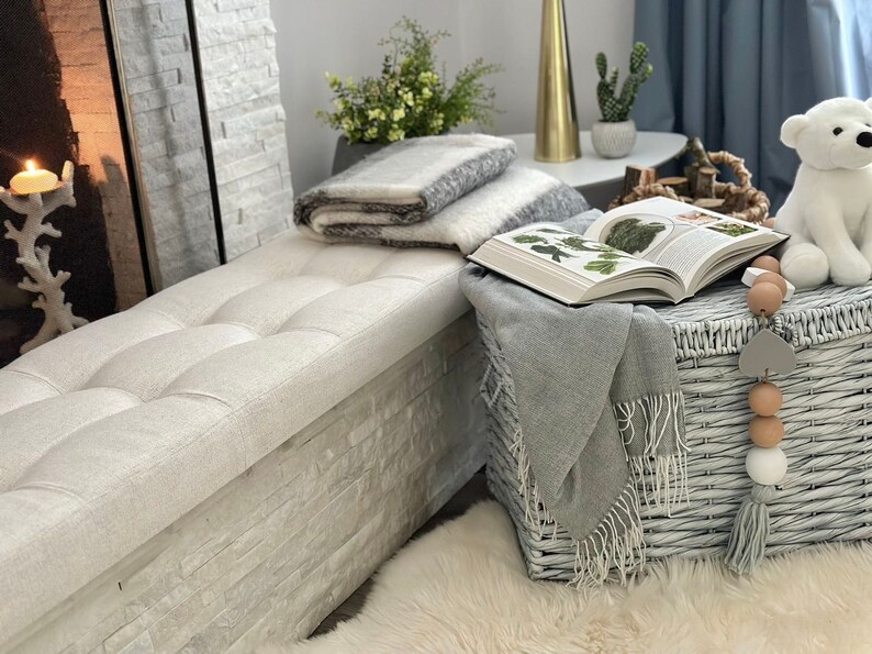 Puede incluir: Una escena interior acogedora con un banco acolchado, una manta a rayas grises y blancas y un libro abierto. Una cesta gris tejida contiene un lanzamiento gris con flecos. Un oso de peluche blanco y cuentas decorativas.