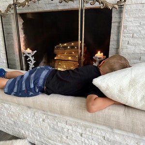 Puede incluir: Una pantalla decorativa para chimenea con una parte superior dorada adornada, un centro de malla negra y una fachada de piedra blanca. Una persona est&aacute; acostada en un banco beige con una almohada blanca.