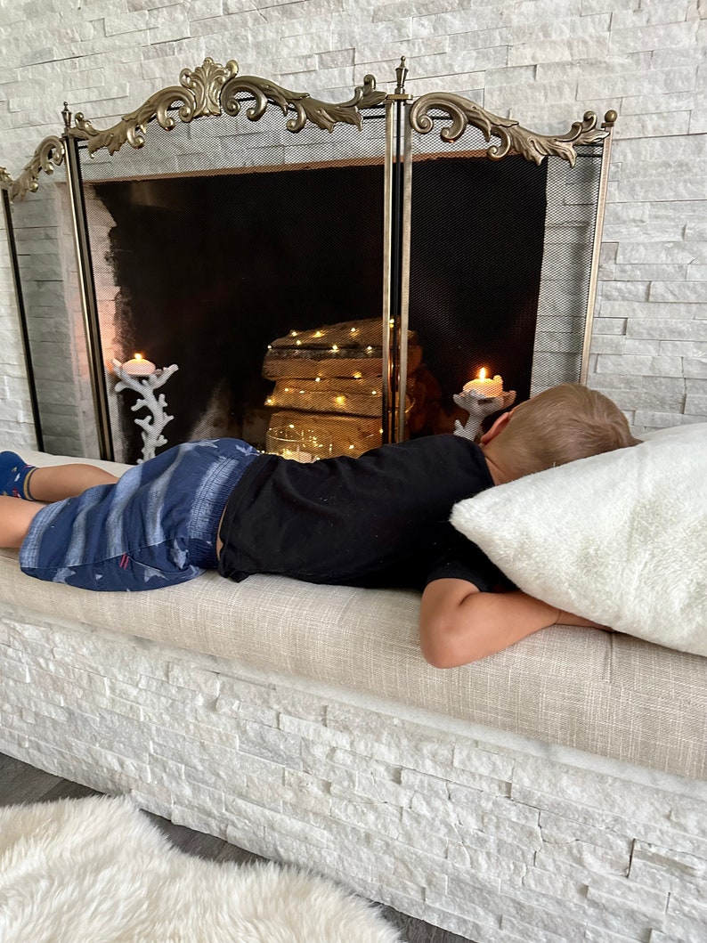 Puede incluir: Una pantalla decorativa para chimenea con una parte superior dorada adornada, un centro de malla negra y una fachada de piedra blanca. Una persona est&aacute; acostada en un banco beige con una almohada blanca.