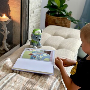 Puede incluir: Un ni&ntilde;o lee un libro con ilustraciones coloridas, acompa&ntilde;ado de un peluche de un personaje de dibujos animados. Una vela encendida en un portavelas decorativo cerca de una chimenea. La escena se desarrolla en un banco de tonos neutros.