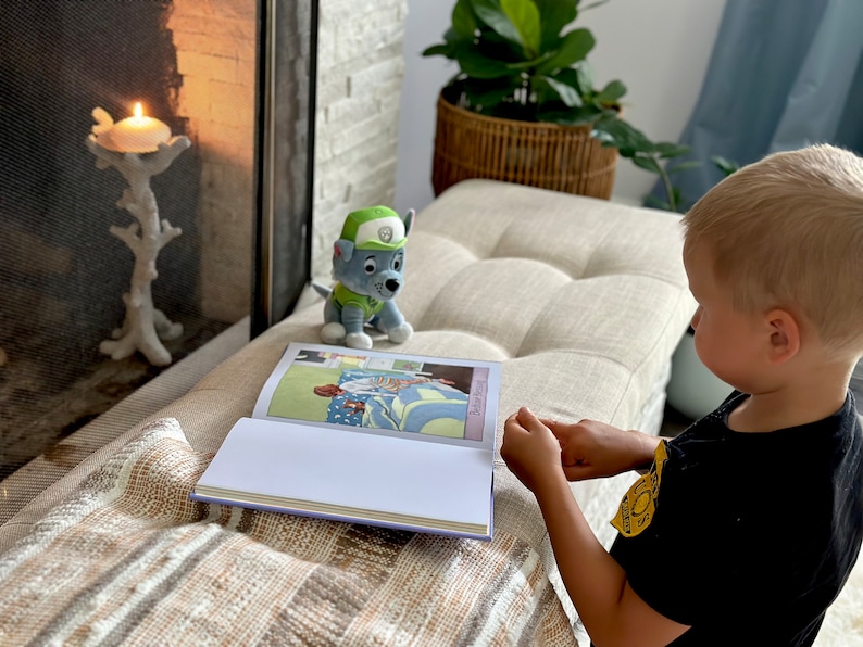 Puede incluir: Un ni&ntilde;o lee un libro con ilustraciones coloridas, acompa&ntilde;ado de un peluche de un personaje de dibujos animados. Una vela encendida en un portavelas decorativo cerca de una chimenea. La escena se desarrolla en un banco de tonos neutros.