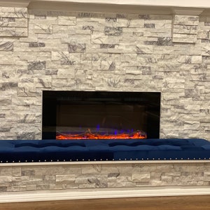 May include: A navy blue tufted bench with a white base and gold stud accents. The bench is in front of a fireplace with a stone facade. The fireplace has a black frame and a simulated flame effect.