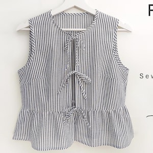 Może przedstawiać: Bezrękawnik w paski z wiązaniem z przodu, wiszący na białym wieszaku. Top ma falisty dół i jest wykonany z tkaniny z pionowymi paskami w kolorze białym i granatowym. Tekst: "Front Tie Top Sewing Pattern 7 Sizes."