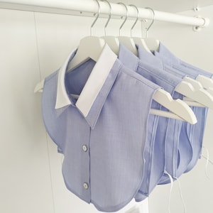 K&ouml;nnte beinhalten: Ein Satz hellblauer Hemden mit wei&szlig;en Kragen, die an wei&szlig;en Kleiderb&uuml;geln in einem Schrank h&auml;ngen. Die Hemden sind gefaltet und haben eine abgerundete Form.