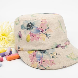 Pode incluir: Um bon&eacute; de lona bege com estampa floral em tons de rosa, azul e amarelo. O bon&eacute; tem aba plana e coroa estruturada.