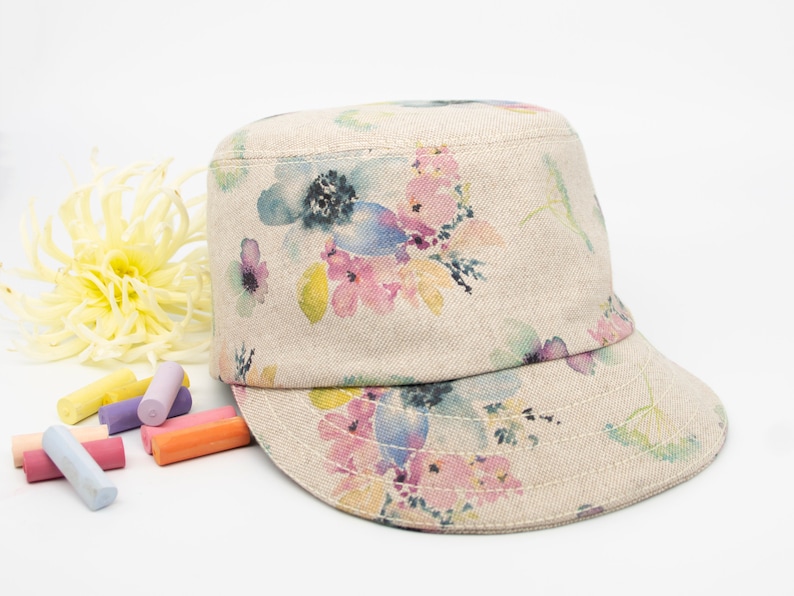 Pode incluir: Um bon&eacute; de lona bege com estampa floral em tons de rosa, azul e amarelo. O bon&eacute; tem aba plana e coroa estruturada.