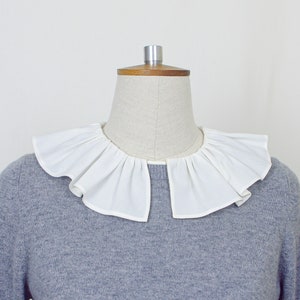 May include: A white ruffled collar on a gray sweater. The collar is made of a soft, lightweight fabric and has a delicate, feminine look.