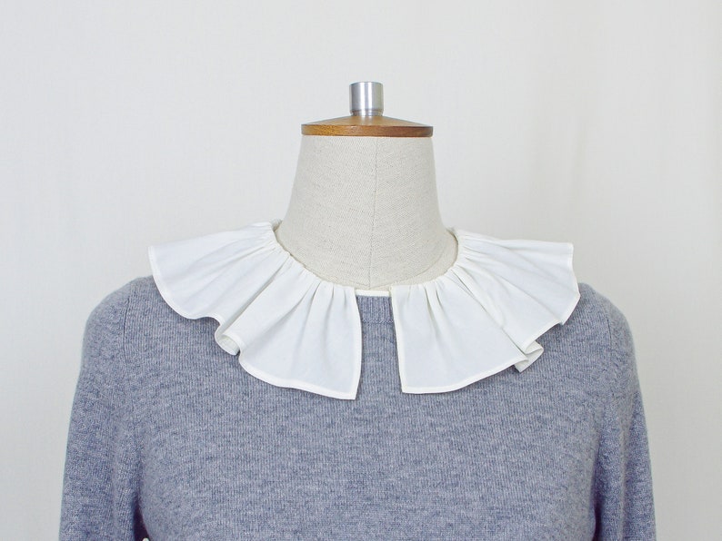 May include: A white ruffled collar on a gray sweater. The collar is made of a soft, lightweight fabric and has a delicate, feminine look.