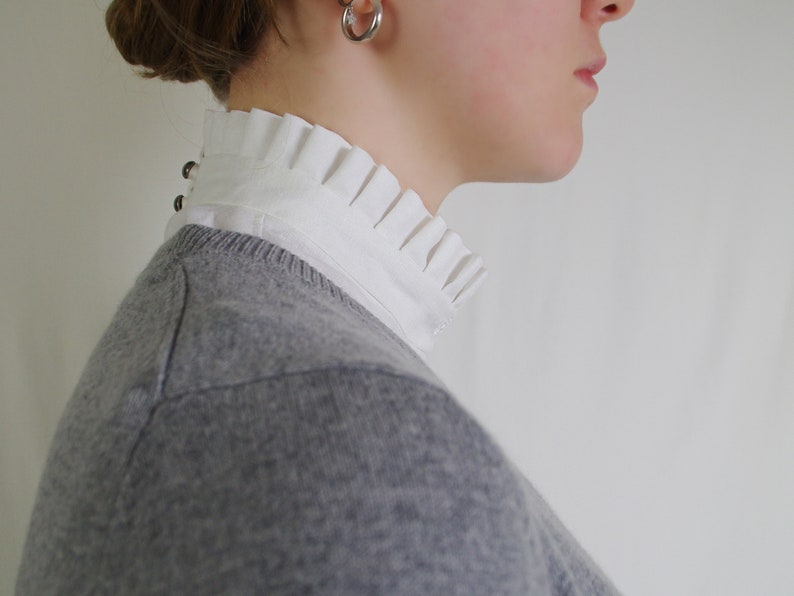 May include: A close-up of a person wearing a gray sweater over a white collared shirt with a ruffled neckline. The shirt has a row of small silver buttons down the back.