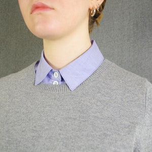 K&ouml;nnte beinhalten: Ein grauer Pullover mit einem hellblauen Hemd mit Kragen darunter. Der Kragen des Hemdes ist &uuml;ber den Halsausschnitt des Pullovers gefaltet.