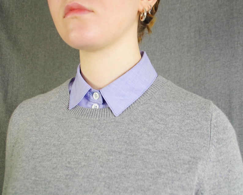 K&ouml;nnte beinhalten: Ein grauer Pullover mit einem hellblauen Hemd mit Kragen darunter. Der Kragen des Hemdes ist &uuml;ber den Halsausschnitt des Pullovers gefaltet.
