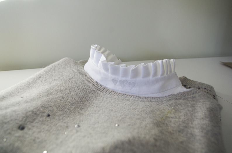 May include: A gray sweater with a white ruffled collar and three embroidered hearts. The sweater has a subtle sparkle.