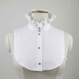 May include: White linen collar with a ruffled edge and silver button closures. The collar is on a white mannequin.