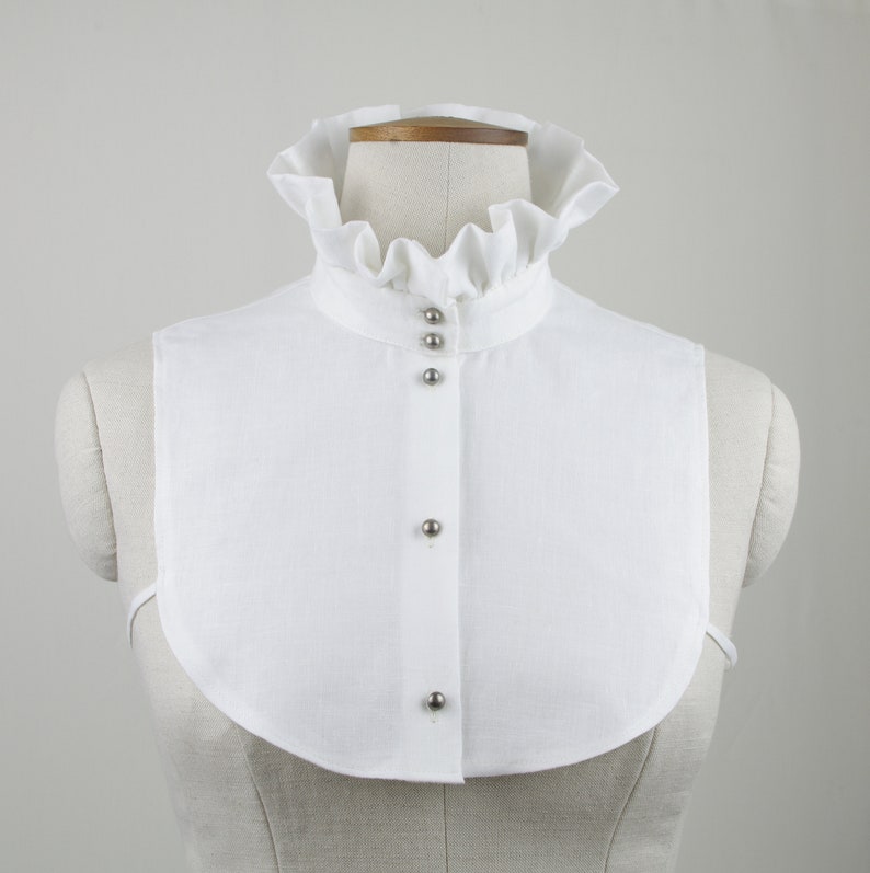 May include: White linen collar with a ruffled edge and silver button closures. The collar is on a white mannequin.