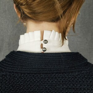 May include: A black knitted sweater with a white ruffled collar and two silver buttons.