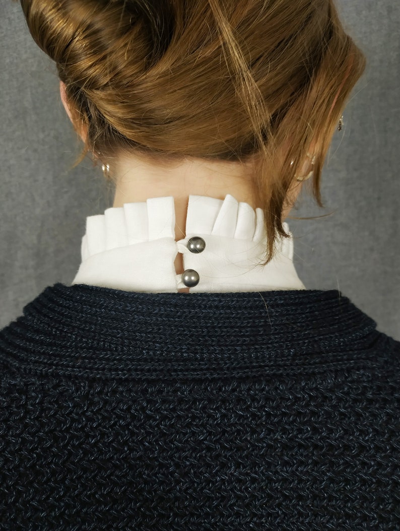 May include: A black knitted sweater with a white ruffled collar and two silver buttons.